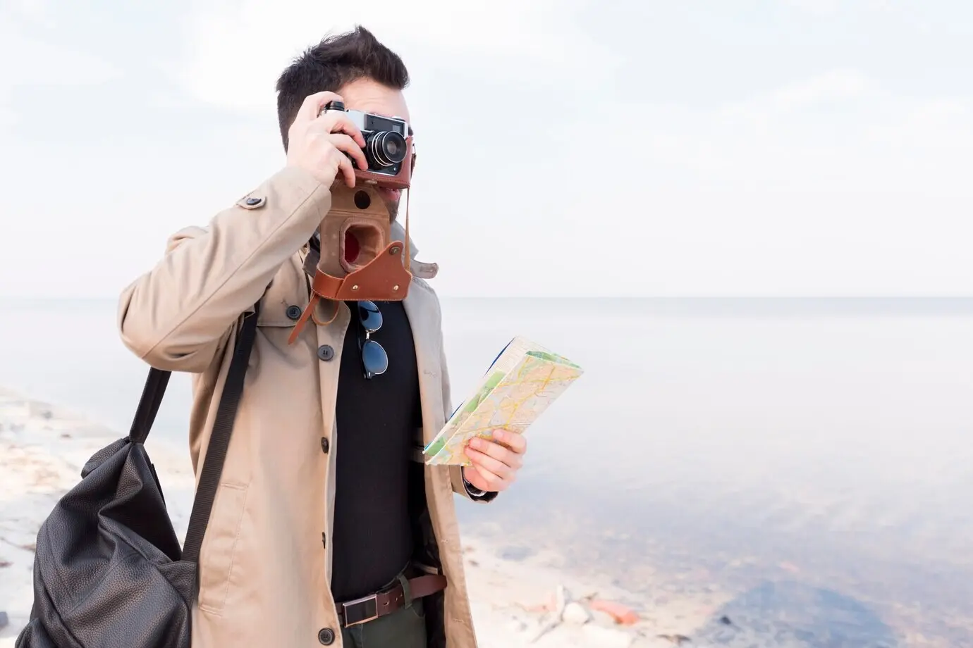 Ein männlicher Reisender hält eine Karte in der Hand und fotografiert am Strand mit einer Kamera.